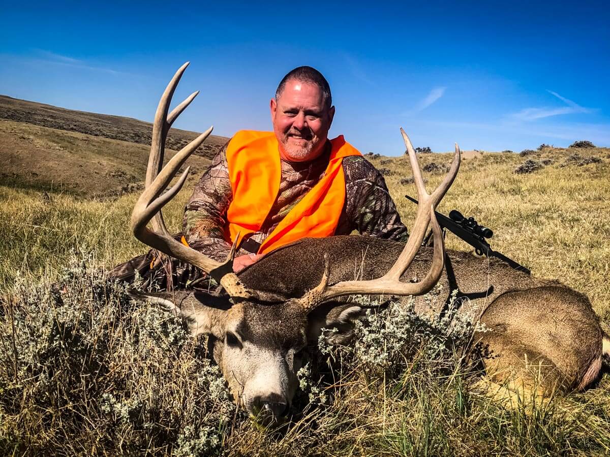 Man in orange vest holding a large deer with prominent antlers in a grassy field.