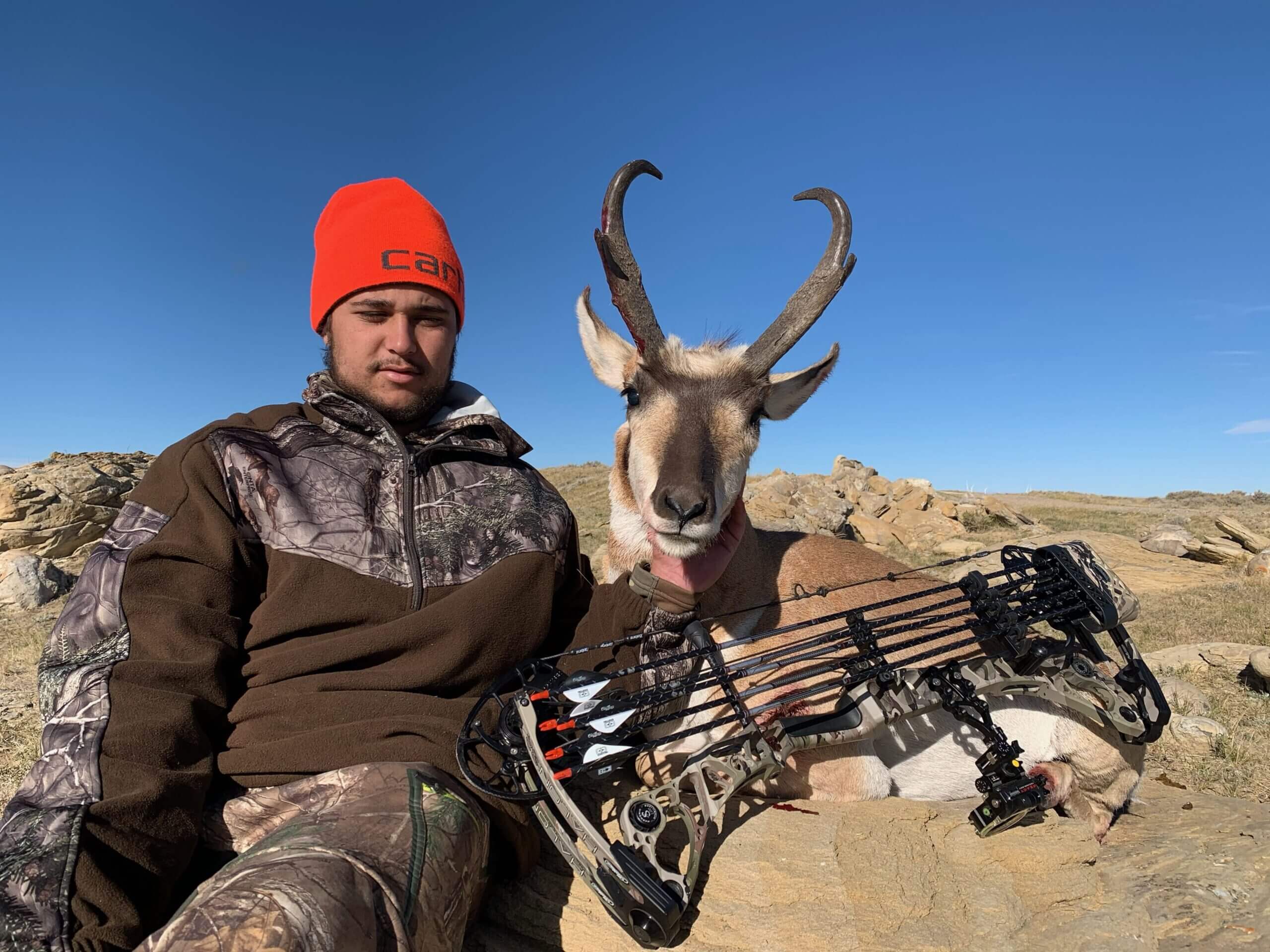 Hunter in camouflage clothing and red beanie with a bow and an antelope on a rocky landscape.