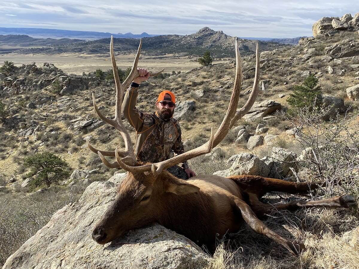 Hunter posing with a large elk in a mountainous landscape