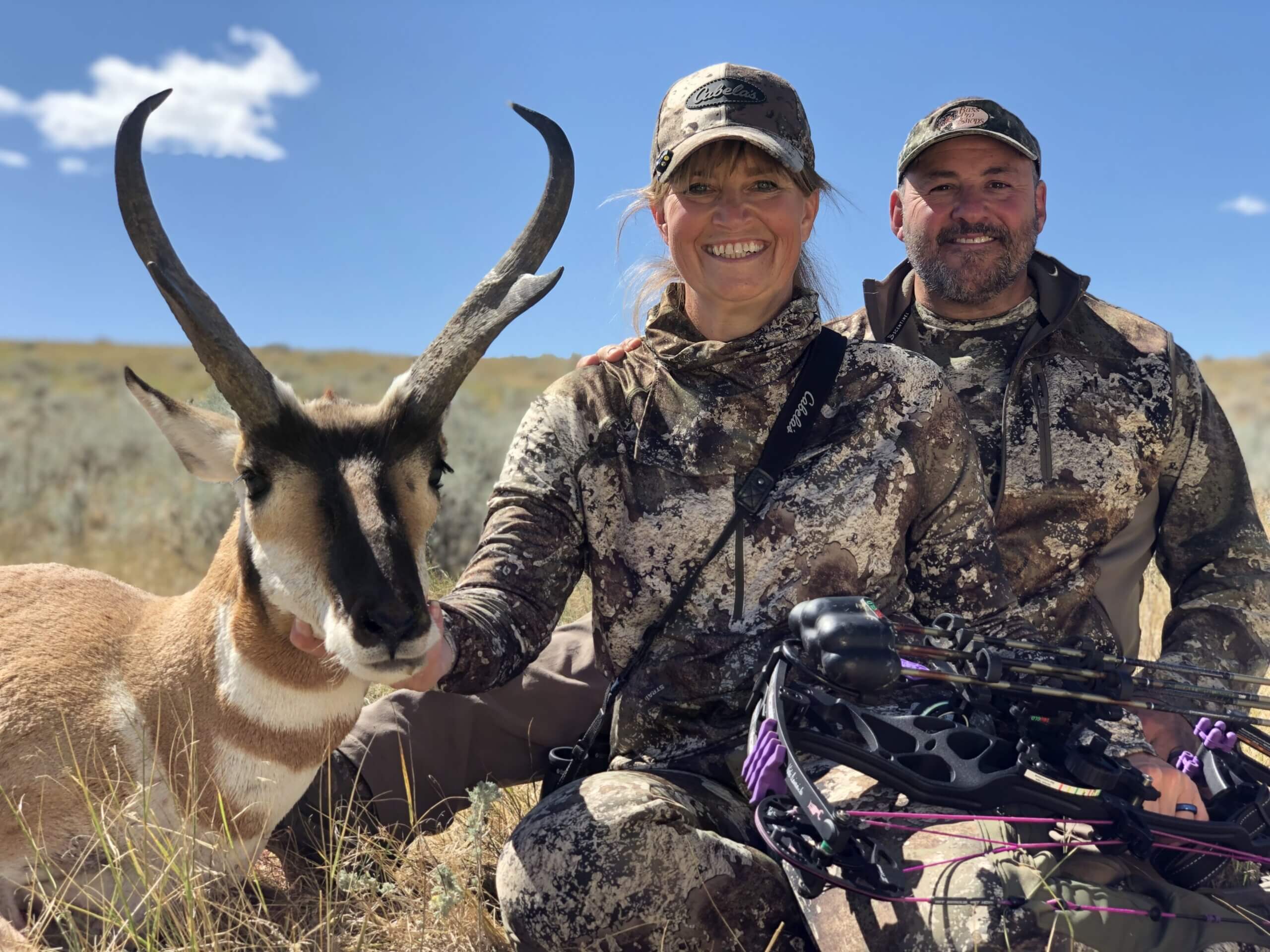 Two hunters in camouflage posing with a bow and a hunted antelope on a grassy field.
