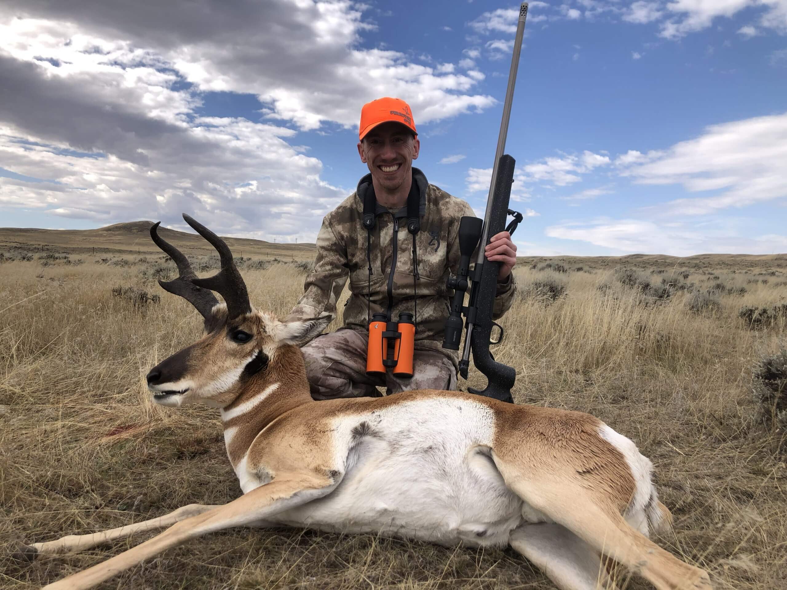 Hunter with a rifle and a harvested antelope in a grassy field under a blue sky.