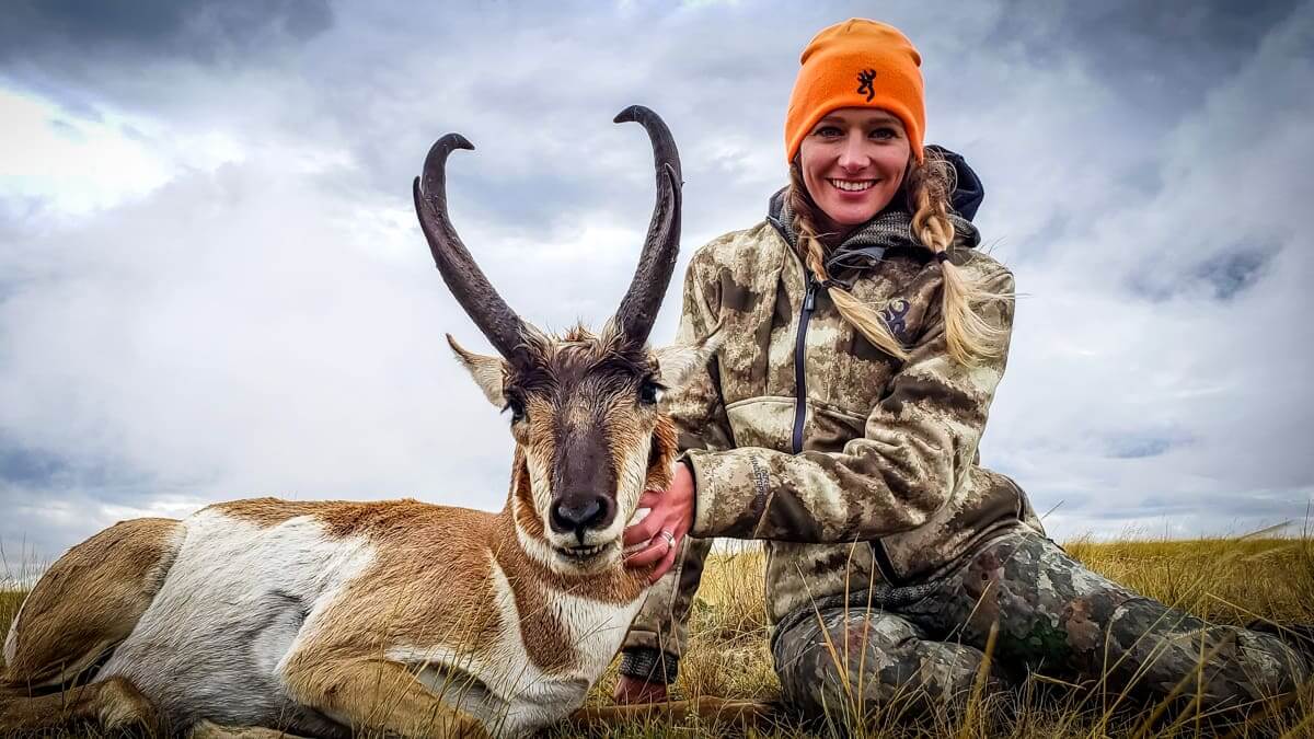 Person in hunting gear with a harvested antelope against a cloudy sky.