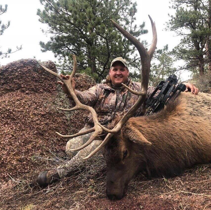 Man in camouflage holding a large elk with a rifle over his shoulder, surrounded by trees.