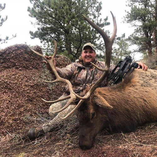 Man in camouflage holding a large elk with a rifle over his shoulder, surrounded by trees.